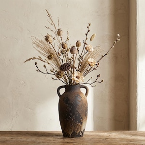 May include: A dark brown, rustic vase with two handles, holding a display of dried flowers and grasses. The vase has a textured, aged look, and the arrangement features beige and tan tones, creating a natural, earthy feel. The vase is on a wooden surface.