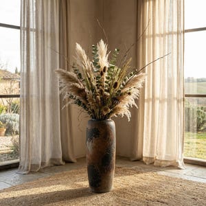 Op de afbeelding: Een hoge, getextureerde bruine vaas met een donker, gevlekt patroon bevat een groot arrangement van gedroogde bloemen en grassen. Het arrangement omvat pampasgras, eucalyptus en andere gedroogde botanische elementen. De vaas staat op een geweven jute vloerkleed.
