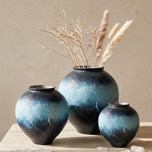 May include: Three spherical ceramic vases in shades of blue and black. The largest vase contains dried grasses and branches. The vases have a gradient effect and are displayed on a light-coloured surface. The vases are of varying sizes.