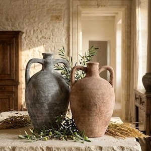 Puede incluir: Dos jarrones grandes y texturizados con asas, uno gris y otro marrón, sobre una superficie de piedra. Ramas de olivo y espigas de trigo dispuestas alrededor de los jarrones. El fondo presenta una pared de piedra y una puerta.