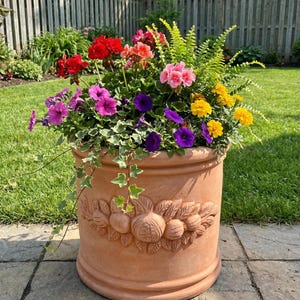 May include: A large terracotta plant pot overflowing with vibrant flowers in shades of red, pink, purple, and yellow, with green foliage. The pot features a decorative fruit and leaf relief. The pot is placed on a stone patio.