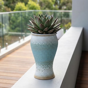 Op de afbeelding: Een keramische plantenbak met een blauw-witte ombre glazuur bevat een groene vetplant. De plantenbak heeft een textuur en een uitlopende vorm. De plant heeft meerdere groene bladeren. De plantenbak staat op een witte richel.