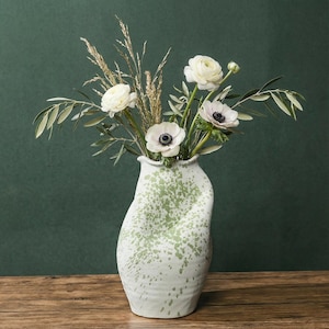 Pode incluir: Um vaso de cerâmica branco com um design de respingos verdes contém um buquê de flores brancas e vegetação. O vaso tem uma forma ondulada única e fica em uma superfície de madeira. As flores incluem anêmonas e ranúnculos.