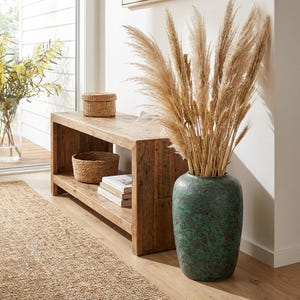 Può includere: Una panca rustica in legno con un ripiano inferiore contiene un cestino intrecciato, libri e una piccola scatola intrecciata. Un grande vaso strutturato, color verde acqua, contiene alte erbe ornamentali essiccate. Sono visibili anche un tappeto in fibra naturale e un vaso di fiori gialli.