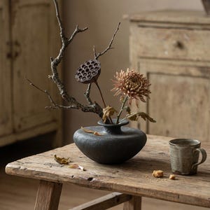 Peut inclure: Une nature morte avec un vase en céramique gris foncé contenant une gousse de lotus séchée, un chrysanthème pêche et une brindille. Une table en bois rustique supporte le vase, une petite tasse et quelques feuilles et miettes séchées. L'arrière-plan comprend des meubles anciens.