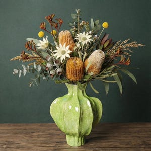 Puede incluir: Un vibrante ramo de flores secas en un jarrón de cerámica verde claro. El arreglo incluye banksias naranjas, margaritas blancas y hojas de eucalipto. El jarrón tiene un diseño acanalado único y está sobre una superficie de madera.
