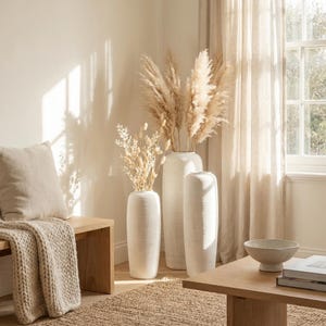Puede incluir: Escena interior de tonos neutros con tres jarrones de cerámica blanca de diferentes alturas, llenos de hierbas ornamentales secas. Un banco de madera con una manta y un cojín beige está a la izquierda. Una mesa de centro de madera y una alfombra están en primer plano.