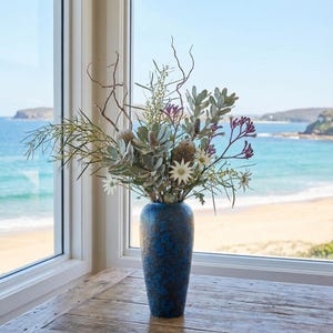 Könnte beinhalten: Eine blaue, strukturierte Vase mit einem Blumenarrangement aus grünen, weißen und lila Blumen und Zweigen. Die Vase steht auf einem Holztisch vor einem Fenster mit Blick auf einen Strand und das Meer.