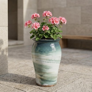 May include: A tall, glazed ceramic planter with a teal and white swirl design, holding a vibrant pink geranium plant. The planter is outdoors, adding a touch of colour to the space. The plant has green leaves and clusters of pink flowers.