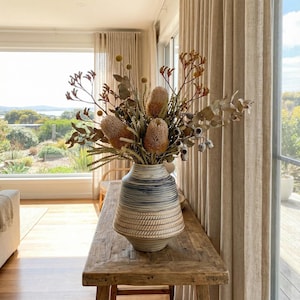 May include: A decorative vase with a blue and beige striped pattern holds a dried floral arrangement. The arrangement includes several large, tan-colored banksia flowers and various dried leaves and seed pods. The vase sits on a rustic wooden table.
