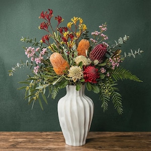 Puede incluir: Un vibrante arreglo floral en un jarrón blanco y acanalado. El ramo presenta flores naranjas, rojas, amarillas y rosas, con follaje verde. El jarrón está sobre una superficie de madera contra un fondo verde oscuro.
