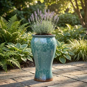 Puede incluir: Una maceta alta de cerámica turquesa, de unos 60 cm de altura, con un esmalte moteado, que contiene una planta de lavanda con flores moradas. La maceta tiene una parte superior acampanada y una base más estrecha con un borde marrón. El fondo presenta follaje verde.