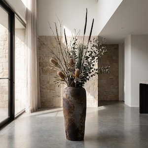 May include: A large, textured brown vase containing dried flowers and foliage. The arrangement features dried eucalyptus and seed pods. The vase is positioned on a polished concrete floor. The vase is approximately 120 cm tall.