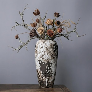 May include: A tall ceramic vase with a white and brown mottled design holds dried flowers and branches. The arrangement includes tan, orange, and brown roses, seed pods, and lichen-covered twigs. The vase sits on a wooden surface.