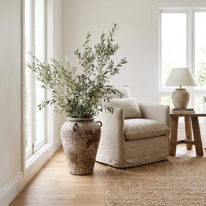 May include: A large, weathered, brown and beige ceramic vase with handles holds a lush arrangement of olive branches. A beige armchair with a matching pillow sits nearby. A small wooden side table holds a lamp with a beige shade.