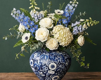 Peut inclure: Un arrangement floral dans un vase à motifs bleu et blanc. Le bouquet comprend des roses blanches, des delphiniums bleus, des anémones blanches et du feuillage vert. Le vase est posé sur une surface en bois.