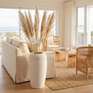 May include: A cream-coloured, textured vase holds dried pampas grass, set in a bright living room. The room includes a beige sofa, wooden furniture, and a natural fibre rug. Large windows provide a view of the beach.