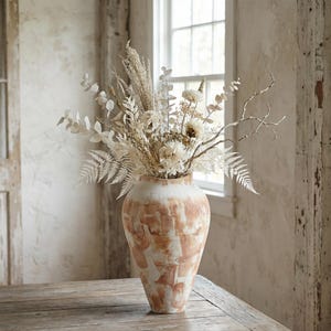 Può includere: Un grande vaso color terracotta con la parte superiore bianca, pieno di fiori e rami secchi. La composizione comprende felci bianche, eucalipto e altri elementi naturali. Il vaso è su un tavolo di legno vicino a una finestra.