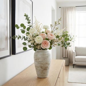 May include: A floral arrangement in a textured, off-white vase. The bouquet features pink and white roses, white filler flowers, and green eucalyptus leaves. The vase sits on a wooden surface, with framed artwork visible in the background.