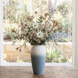 Könnte beinhalten: Ein Blumenstrauß aus weißen und rosa Blüten mit grünem Laub in einer zweifarbigen Vase. Die Vase wechselt von Weiß zu einem gedämpften Blau. Die Anordnung steht auf einem Holztisch vor einem Fenster.