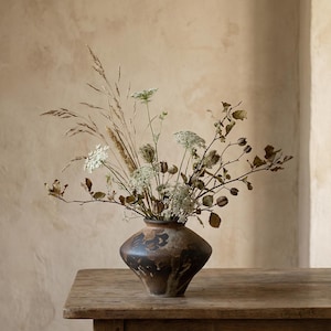 Puede incluir: Un jarrón marrón rústico con flores y hierbas secas. El arreglo presenta tonos blancos, marrones y beige, creando una estética natural. El jarrón está sobre una mesa de madera. El jarrón tiene una forma única.