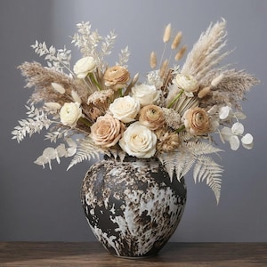 May include: A large floral arrangement in a decorative vase. The bouquet features cream and tan roses, pampas grass, and other dried flowers. The vase is a dark brown with a white splatter design. The arrangement is on a wooden surface.