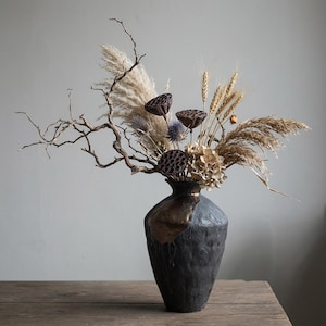May include: A dark gray vase with a gold accent holds a dried floral arrangement. The arrangement includes pampas grass, wheat, seed pods, and dried hydrangeas. The vase sits on a wooden surface, creating a rustic and natural aesthetic.