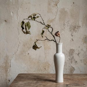 May include: A tall, white ceramic vase contains a dried branch with brown leaves and a dried fruit. The vase is placed on a wooden surface, set against a textured, off-white wall.
