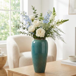 Könnte beinhalten: Eine hohe, türkisfarbene Keramikvase mit einem Blumenarrangement. Der Strauß enthält weiße Rosen, blaue Rittersporne und grünen Eukalyptus. Die Vase steht auf einem Holztisch, im Hintergrund ein Stuhl in neutralen Tönen und ein Fenster.