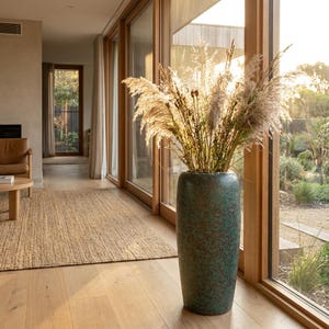 Puede incluir: Un jarrón alto y cilíndrico con un acabado moteado en verde azulado y marrón, lleno de hierbas ornamentales secas. El jarrón está cerca de una ventana grande, con luz natural que ilumina el interior y el jardín exterior.