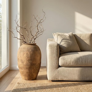May include: A large, textured, brown ceramic vase filled with dried branches. The vase has vertical carved lines and sits on a wooden floor next to a beige sofa. A woven rug is in the foreground, and a window is on the left.