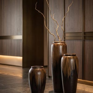 May include: Three cylindrical ceramic floor vases with a dark brown and tan drip glaze finish. The vases vary in height, with the tallest one containing several thin, twisted, light-coloured decorative branches extending upwards from the opening.