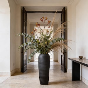 Könnte beinhalten: Eine hohe, schwarze, zylindrische Vase gefüllt mit einem Arrangement aus getrockneten Gräsern, Eukalyptus und bunten Blumen. Die Vase steht in einem Flur mit offenen dunklen Holztüren und einem Marmorboden.