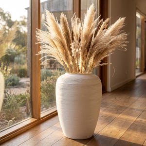 Può includere: Un grande vaso bianco sporco strutturato pieno di erba della pampa essiccata. Il vaso è cilindrico con una base ampia e un collo leggermente più stretto. Le piume di erba della pampa sono beige e marroni, creando una palette di colori naturali. Il vaso è vicino a una finestra.