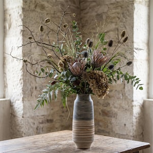 May include: A vase filled with a vibrant arrangement of dried flowers and foliage. The vase features a two-tone design in grey and beige. The bouquet includes protea, thistles, and eucalyptus, creating a natural, rustic aesthetic. The vase is on a wooden table.