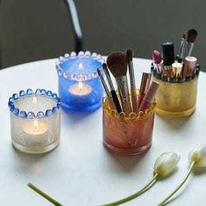 Könnte beinhalten: Dekorative Glashalter in verschiedenen Farben. Zwei Halter enthalten brennende Kerzen, während zwei weitere Make-up-Pinsel und Lippenstifte enthalten. Die Halter haben einen Perlenrand und stehen auf einer weißen Oberfläche.