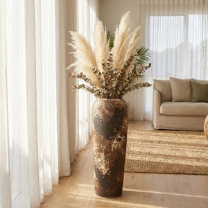 Puede incluir: Un jarrón alto y marrón rústico lleno de hierba de la pampa seca y eucalipto. El jarrón tiene un acabado moteado y texturizado y se encuentra frente a una ventana con cortinas blancas transparentes. El arreglo añade un toque natural y bohemio.