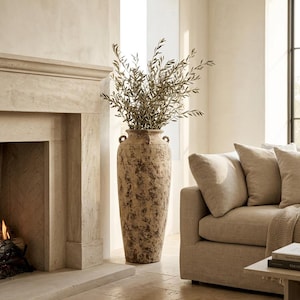 Op de afbeelding: Een hoge, op de vloer staande terracotta vaas met een verweerde, gevlekte beige en bruine afwerking, voorzien van twee kleine handvatten bij de rand. De vaas is gevuld met lange, slanke takken met kleine, smalle groene bladeren.