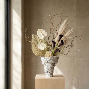 May include: A contemporary floral display in a speckled vase. The arrangement includes white calla lilies, dark purple calla lilies, dried palm leaves, pampas grass, and slender branches. The vase is placed on a light wood block.