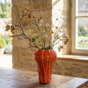 May include: An orange ceramic vase with a ribbed design, filled with dried flowers and branches. The vase sits on a wooden table. The vase is approximately 30 cm tall, with a stone wall and window in the background.