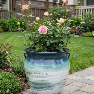 May include: A large, ceramic garden pot with a blue and green ombre glaze, filled with a rose bush. The roses are a soft pink color, with green leaves and stems. The pot has two handles and sits on a brick patio.