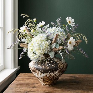 May include: A decorative vase with a floral arrangement. The vase is brown and white, with a speckled design. The bouquet includes white hydrangeas, cosmos, and greenery, with driftwood accents. The vase is on a wooden table.