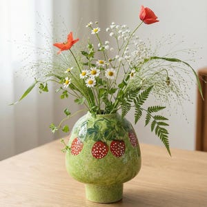 Puede incluir: Un jarrón de cerámica verde con diseño de fresas, que contiene un ramo de flores silvestres. El jarrón tiene forma bulbosa y base de pedestal. Las flores incluyen amapolas rojas, margaritas blancas y follaje verde.