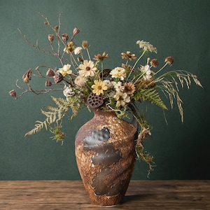 May include: A rustic, brown ceramic vase with a textured surface, filled with a dried floral arrangement. The arrangement includes beige daisies, seed pods, and fern fronds, creating a natural, earthy aesthetic. The vase sits on a wooden surface.