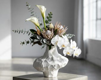 May include: A white textured ceramic vase filled with a floral arrangement. The arrangement includes white calla lilies, orchids, and a protea flower, with green and silver foliage. The vase sits on a gray concrete block.