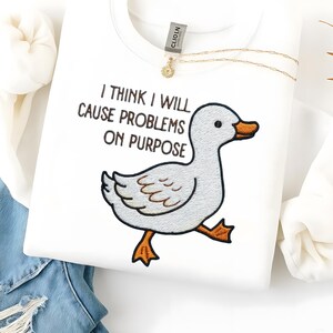 May include: White sweatshirt with the embroidered phrase "I THINK I WILL CAUSE PROBLEMS ON PURPOSE" and a cartoon duck. The duck has gray feathers, an orange beak and feet. A gold necklace is draped across the sweatshirt.