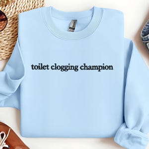 May include: A light blue sweatshirt with the text "toilet clogging champion" in black. The sweatshirt has a crew neck and long sleeves. The Gildan brand tag is visible on the inside of the collar.