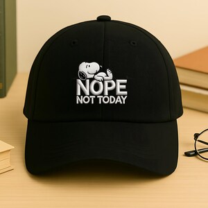 May include: Black baseball cap featuring a white embroidered Snoopy graphic with the text "NOPE NOT TODAY." The cap has a curved brim and a button on top. A fun and casual accessory.