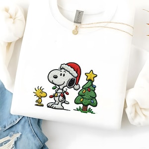 以下が含まれることがあります： サンタ帽をかぶったスヌーピーとクリスマスツリーの刺繍が施された白いスウェットシャツ。クリスマスライトで飾られています。小さな黄色い鳥もデザインに含まれています。金のネックレスも見えます。