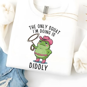 Może przedstawiać: Biała bluza z kapturem z kreskówkową żabą w różowym kapeluszu kowbojskim i butach, trzymającą lasso. Tekst "THE ONLY SQUAT I'M DOING IS DIDDLY" jest wydrukowany nad i pod ilustracją żaby. Złoty naszyjnik wisi na koszuli.